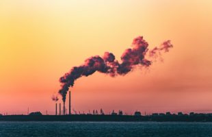 Rookpluimen boven een industrieterrein onder een gekleurde avondlucht, symbool voor de uitstoot van fossiele brandstoffen in de energiemarkt.