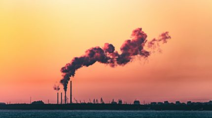 Rookpluimen boven een industrieterrein onder een gekleurde avondlucht, symbool voor de uitstoot van fossiele brandstoffen in de energiemarkt.