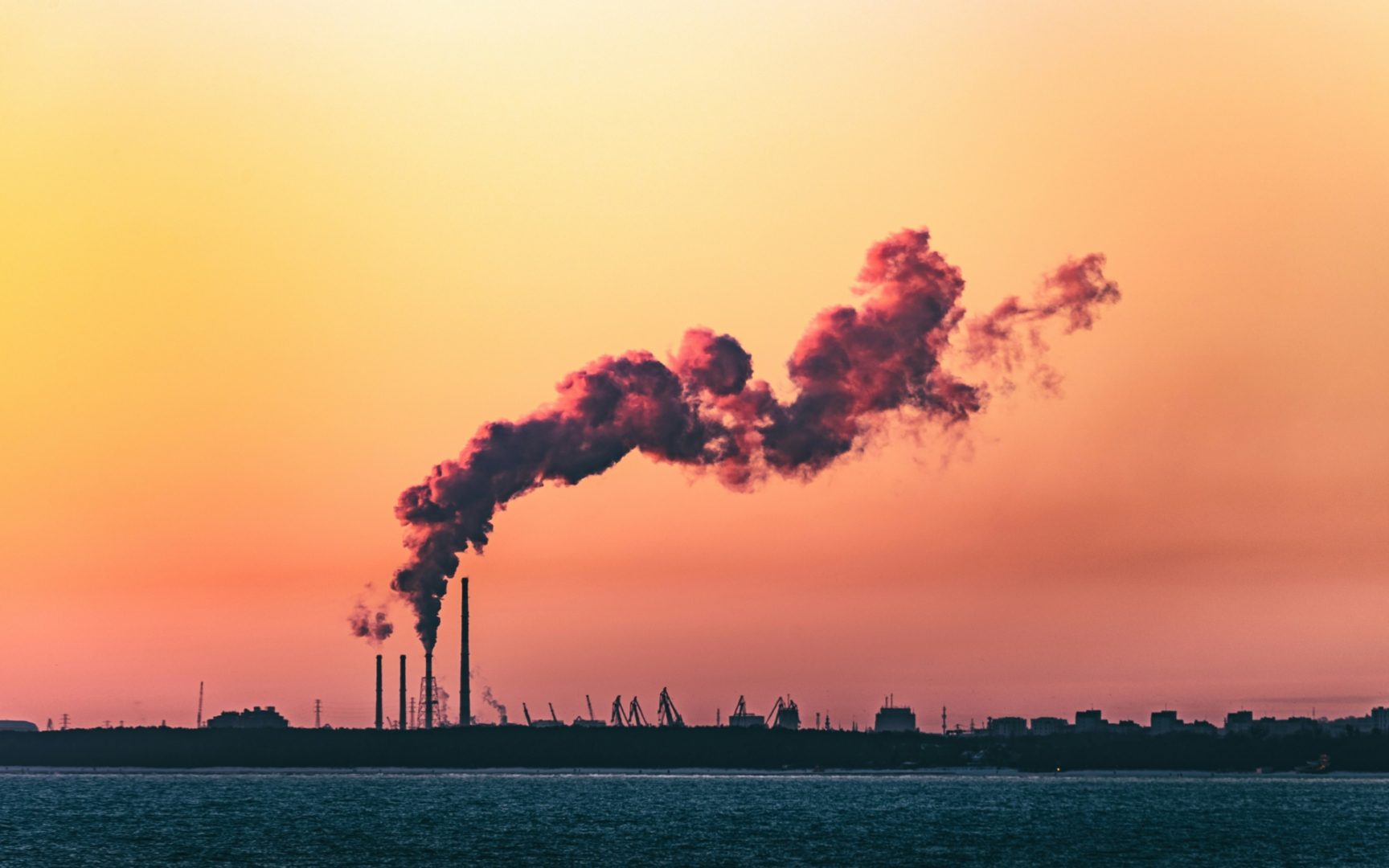 Rookpluimen boven een industrieterrein onder een gekleurde avondlucht, symbool voor de uitstoot van fossiele brandstoffen in de energiemarkt.